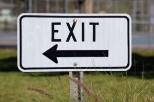 Exit sign with left-pointing arrow in a grassy outdoor setting.