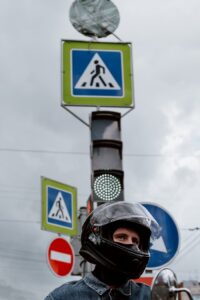 A motorcyclist wearing a helmet waits at a traffic light beside street signs on a cloudy day.