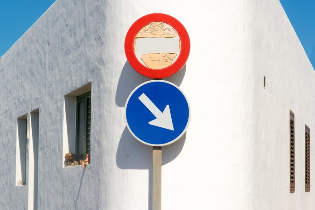 pexels-photo-8906892 Blue and red road signs against a bright white wall under clear blue sky. Minimalist composition.